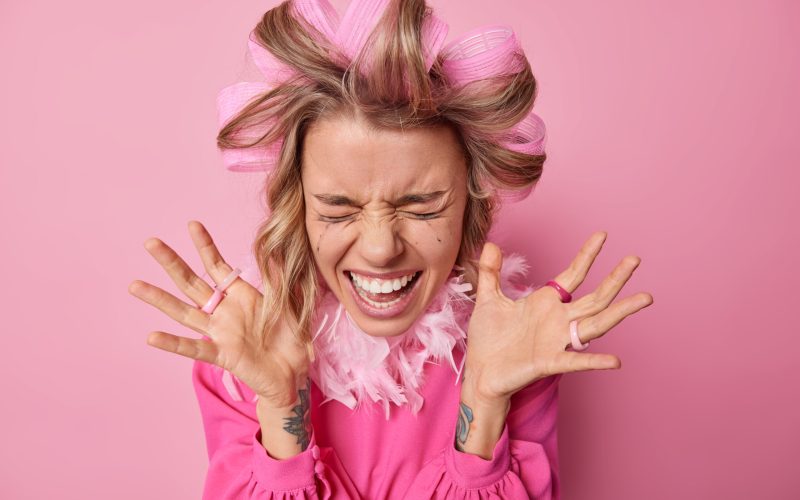 Happy emotional young woman keeps palms raised shouts loudly reacts on something amazing wears hair rollers for making curly hairstyle gets ready for date isolated over pink studio background